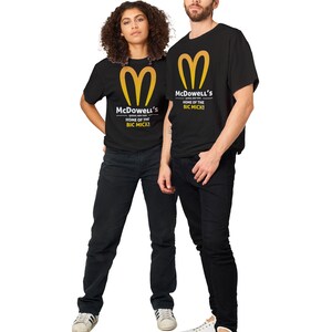 May include: Two people wearing black t-shirts with a gold logo and text that reads "McDowell's HOME OF THE BIG MICK!". The shirts are paired with black jeans and white sneakers. The logo is a stylized golden arch.