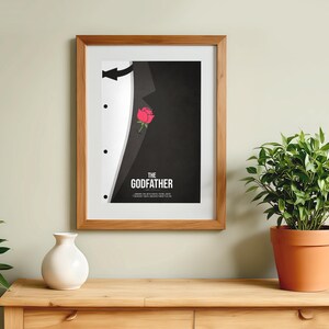 May include: Framed art print featuring a stylized tuxedo design with a red rose and the text "THE GODFATHER" in white. The artwork is in a wooden frame, displayed on a wooden surface, with a white vase and potted plant nearby.