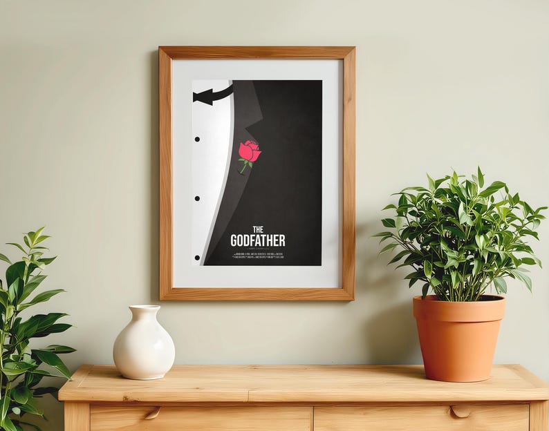 May include: Framed art print featuring a stylized tuxedo design with a red rose and the text "THE GODFATHER" in white. The artwork is in a wooden frame, displayed on a wooden surface, with a white vase and potted plant nearby.