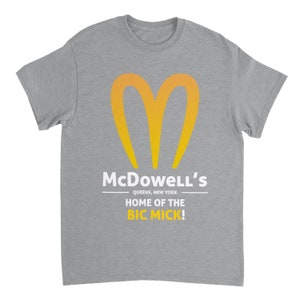 May include: A heather gray t-shirt featuring a stylized yellow and orange heart-shaped logo. Below the logo, the text reads "McDowell's QUEENS, NEW YORK HOME OF THE BIC MICK!" in white.
