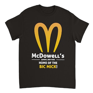 May include: Black t-shirt featuring a yellow and orange heart-shaped logo. The text "McDowell's" is above "QUEENS, NEW YORK" and "HOME OF THE BIC MICK!" in white.