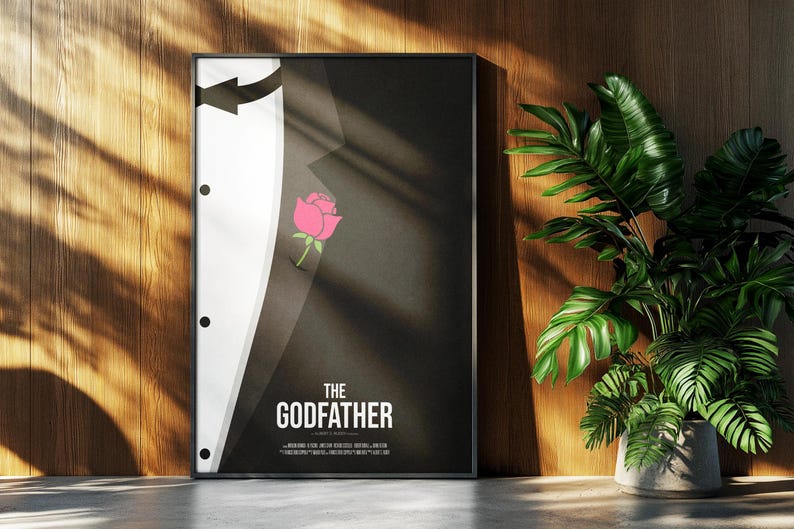 May include: Framed poster featuring a minimalist design inspired by "The Godfather" film. The artwork depicts a black suit with a white shirt, a pink rose in the pocket, and the film's title in white text. The poster is displayed against a wooden wall.