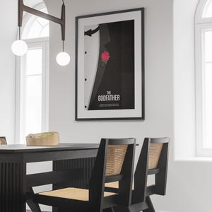 May include: Framed poster featuring a minimalist design inspired by "The Godfather." The artwork depicts a black suit with a red rose in the pocket and the film's title in white font. The poster is displayed in a dining room setting.