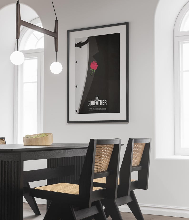 May include: Framed poster featuring a minimalist design inspired by "The Godfather." The artwork depicts a black suit with a red rose in the pocket and the film's title in white font. The poster is displayed in a dining room setting.