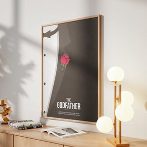 May include: Framed poster featuring a minimalist design inspired by "The Godfather" film. The artwork depicts a black suit with a white shirt and a red rose. The poster is mounted on a light wood frame and hangs on a white wall.