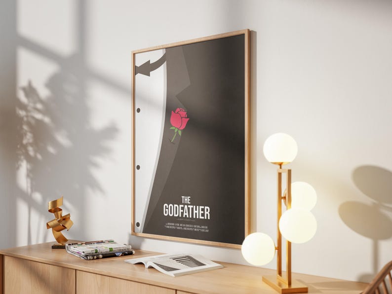 May include: Framed poster featuring a minimalist design inspired by "The Godfather" film. The artwork depicts a black suit with a white shirt and a red rose. The poster is mounted on a light wood frame and hangs on a white wall.
