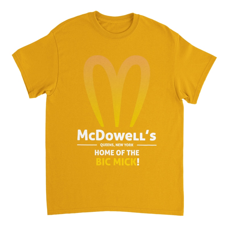 May include: Mustard yellow t-shirt with a stylized, gradient heart-shaped logo. The text "McDowell's" is above "QUEENS, NEW YORK" and "HOME OF THE BIC MICK!" in white. The shirt is a short-sleeved, crew neck style.
