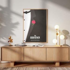 May include: Framed poster featuring a minimalist design from "The Godfather" movie. The artwork shows a black suit with a white shirt and a red rose. The title "THE GODFATHER" is displayed in white text.