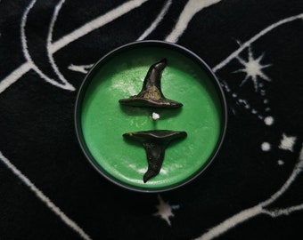 May include: Green candle with two black witch hat toppers. The candle is in a glass jar with a black lid.