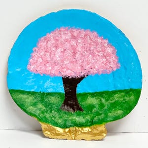 May include: A circular, textured decorative object featuring a hand-painted scene of a tree with a dark brown trunk and a full, textured pink canopy. The tree is set against a bright blue sky and green grass, with a metallic gold base at the bottom.