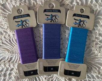 May include: Three spools of 4mm River Silks ribbon in purple, blue, and turquoise. Each spool is labelled with the length in metres and yards, the material, and the country of origin.