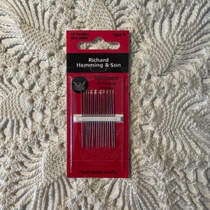 May include: A red package of Richard Hemming & Son Milliners needles, size 9, with the text "Finest Quality Needles". The package contains 16 needles. The background is a white, textured, crocheted surface.