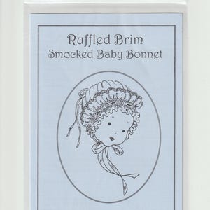 May include: A light blue paper pattern insert for a Ruffled Brim Smocked Baby Bonnet by Jeannie Baumeister, enclosed in a clear plastic bag. The insert features a black line drawing of a baby wearing a bonnet with a ruffled brim and ribbon ties.
