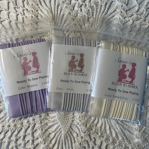 May include: Three packages of ready-to-sew piping in wisteria, silver, and natural ecru. Each package has a label with the text "Susie's Ready To Smock" and the color name. The wisteria and silver packages contain 3 yards of piping.