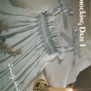 Puede incluir: Un vestido smockado azul claro con ribete de encaje blanco y lazos de cinta azul claro. El vestido se muestra sobre una superficie blanca con un candelabro de latón y una vela encendida. El texto "Smocking Daze I" es visible.
