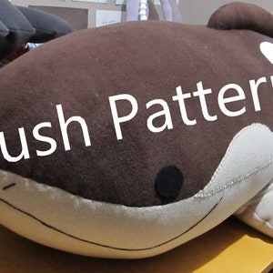 May include: A large, plush whale toy in shades of brown and cream, with the text "Plush Patterns" overlaid. The toy has a rounded shape with fins and a stitched mouth. The plush toy is on a table.