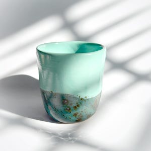May include: A handmade ceramic cup with a smooth, mint green upper half and a textured, teal and gray lower half. The cup has a rounded shape and a glossy finish, perfect for coffee or tea.