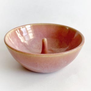 May include: A pink ceramic ring holder with a small, raised center post. The holder has a glossy finish and a subtle speckled pattern.