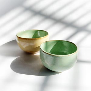 May include: Two ceramic bowls, one light yellow and the other light green, sit side-by-side on a white surface. The bowls have a rounded shape with a slightly textured appearance and a brown rim. The bowls are lit by natural light.