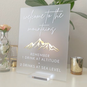 May include: A frosted acrylic sign with white text that reads "Welcome to the mountains" and a gold mountain silhouette. The sign also includes the text "Remember 1 drink at altitude = 2 drinks at sea level".