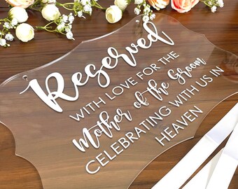 May include: A clear acrylic sign with the text "Reserved with love for the mother of the groom celebrating with us in heaven". The sign has a decorative border and is attached to three white ribbons.
