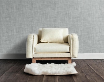 May include: A white armchair with a white cushion sits on a wooden floor in front of a gray textured wall. A white fluffy rug is in front of the chair.