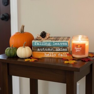 May include: A stack of three decorative wooden blocks painted teal, dark brown, and orange with the text falling leaves, autumn skies, and hello fall. Beside the blocks are three pumpkins, a lit candle labeled Harvest Spice, and autumn leaves.