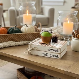 May include: Two stacked white wooden blocks tied with twine, featuring the text "sweet" and "fall treat" in black font. A green caramel apple with a buffalo check bow sits on top, alongside a small pinecone, creating a decorative display.