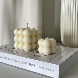 May include: Two ivory bubble cube candles with wicks, resting on a book. The candles have a rounded, textured design. The book reads "that will CHANGE the way YOU THINK". Decorative home decor.