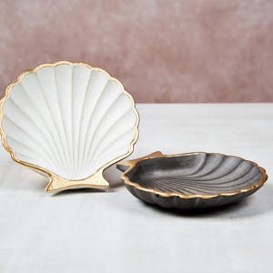 Op de afbeelding: Twee decoratieve keramische schelpen schalen. Eén is wit met gouden rand en de andere is zwart met gouden rand.