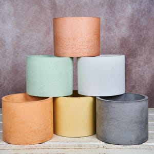 Könnte beinhalten: Eine Sammlung zylindrischer Pflanzgefäße in verschiedenen Farben, darunter Pfirsich, Mintgrün, Weiß, Gelb, Orange und Grau. Die Pflanzgefäße sind pyramidenförmig angeordnet und zeigen ihr schlichtes, modernes Design. Die Pflanzgefäße sind aus Beton.
