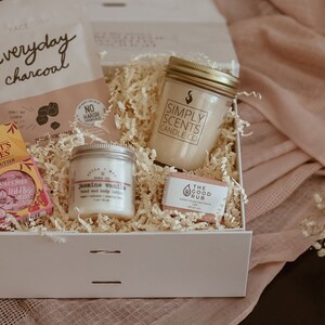 May include: A white gift box filled with self-care items, including a candle, soap, lip balm, and face mask. The box is surrounded by white tissue paper and a pink fabric.