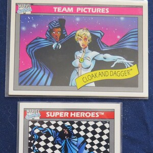 May include: Two trading cards featuring the Marvel Comics characters Cloak and Dagger. The first card shows Cloak and Dagger together, with Dagger in a white costume and Cloak in a dark blue cape. The second card shows Cloak alone, in a blue cape, against a black and white checkered background. Both cards have the Marvel Comics logo and the text "SUPER HEROES TM".
