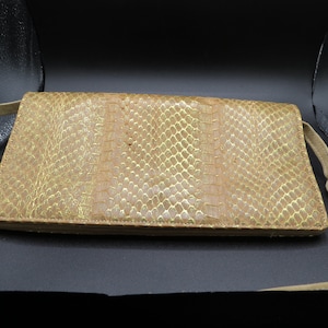May include: A tan clutch bag with a snakeskin pattern and a gold metallic sheen. The rectangular bag has a long, thin strap and a fold-over flap. The bag is made of a textured material.