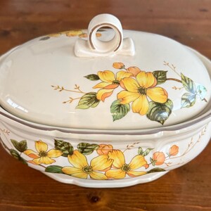 Puede incluir: Una cacerola ovalada de cerámica blanca con tapa. La cacerola tiene un borde marrón y un diseño floral con flores amarillas y verdes.