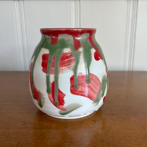 Puede incluir: Un jarrón de cerámica con base blanca, adornado con pinceladas y goteos rojos y verdes. El borde está pintado de rojo. El jarrón tiene forma redondeada y es adecuado para exhibir flores o como pieza decorativa.
