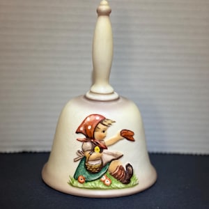 May include: Cream-colored ceramic bell with a handle. The bell features a relief of a girl in a red polka-dot scarf, holding a basket and a loaf of bread. The girl is sitting on a patch of green grass with orange flowers.