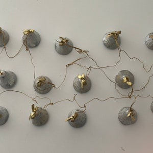 Könnte beinhalten: Eine Lichterkette mit silberfarbenen, muschelförmigen Elementen und goldenen Akzenten. Die Lichter sind durch dünne, goldfarbene Drähte verbunden, die ein einzigartiges Lichtdisplay bilden. Die Lichter sind auf einer weißen Oberfläche montiert.