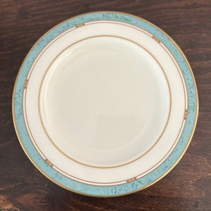 May include: A white ceramic plate with a blue and gold rim. The rim has a decorative pattern of small dots and lines.