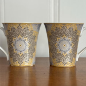 May include: Two white ceramic mugs with gold and silver intricate designs. The mugs have a handle on the right side.
