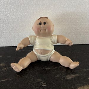 Może przedstawiać: Mała figurka siedzącego dziecka z okrągłą głową, jasną skórą i brązowymi włosami namalowanymi na czubku. Figurka ma na sobie prostą, kremową koszulkę z krótkim rękawem i pasujące majteczki, z neutralnym wyrazem twarzy i ciemnymi oczami.