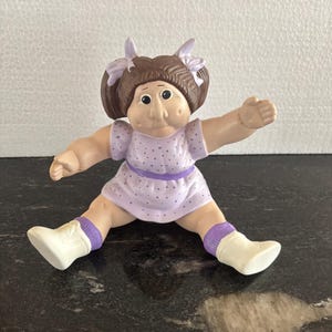 May include: A small plastic doll with brown hair styled in pigtails with light purple bows. The doll wears a light purple dress with a darker purple polka dot pattern and a solid purple waistband, paired with white shoes and purple socks.