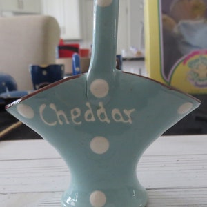 May include: A light blue ceramic cheese dish with white polka dots and the word "Cheddar" written in white. The dish has a handle and a wide, shallow bowl.