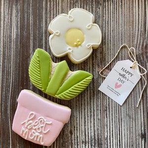May include: A set of three decorated sugar cookies. The cookies are shaped like a flower, a plant, and a pot. The pot is pink and says "best mom". The flower is white with a yellow center. The plant is green. The cookies are on a wooden background. The tag says "Happy Mother's Day" and "Just a little something to sweeten your day."