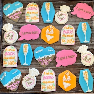 May include: A collection of decorated sugar cookies in various shapes, including hearts, speech bubbles, and hexagons. The cookies feature text like "I got a text!" and "A hot new bride has entered the Villa."