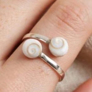 Puede incluir: Un anillo abierto de tono plateado que presenta dos pequeñas conchas de opérculo blancas con patrón en espiral en cada extremo de la banda. El anillo está diseñado con un acabado metálico pulido que se curva alrededor del dedo, mostrando los detalles.