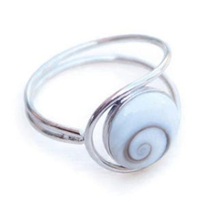Op de afbeelding: Een zilverkleurige metalen ring met een ronde, witte en lichtbruine spiraalvormige schelpinleg. De ringband is gemaakt van een dunne, doorlopende draad die rond het centrale schelpelement loopt, wat zorgt voor een open en licht ontwerp.