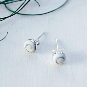 Op de afbeelding: Een paar oorknopjes met kleine, ronde, witte shiva-schelp cabochons met een natuurlijk spiraalpatroon, elk gezet in een gepolijste zilverkleurige metalen zetting met een standaard stift en een bijpassende zilverkleurige vlindersluiting.