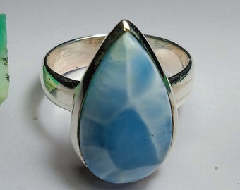 May include: A silver ring featuring a pear-shaped, light blue gemstone with white veining. The gemstone is set in a silver bezel, and the ring band is a simple, polished silver. The ring is displayed on a white surface.