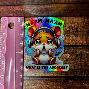 Pode incluir: Um adesivo hologr&aacute;fico com um hamster de desenho animado usando fones de ouvido e uma express&atilde;o surpresa. O texto "MA'AM, MA'AM WHAT IS THE ADDRESS?" est&aacute; impresso abaixo do hamster.